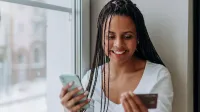 Mujer sonriente con smartphone y tarjeta de crédito para compras en línea en interiores con luz natural.