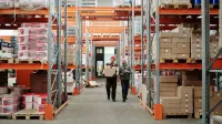 Two workers handle a package in a spacious warehouse surrounded by shelves stocked with boxes and products.