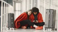 Feliz pareja sentada en el piso del aeropuerto con equipaje, compartiendo un momento con smartphone.
