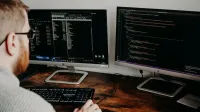 Desarrollador de software que trabaja en código con monitores duales en un entorno de oficina en el hogar.
