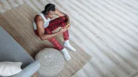 A man in plaid pajamas sits on the floor wearing headphones, using his smartphone in a cozy living room.
