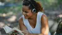 Cheerful young Hispanic female in white tank top listening to music via headphones and reading book while sitting on chair in park