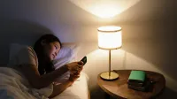A woman relaxes in bed, using her smartphone beside a lit table lamp in a cozy room.