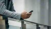 Hombre de negocios negro anónimo de cultivo en traje formal apoyado en la cerca y usando el teléfono a la luz del día suave