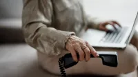 Una persona se sienta en un sofá escribiendo en una computadora portátil y sosteniendo un receptor de teléfono negro con la mano derecha.