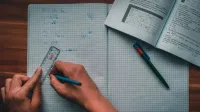 Una persona escribe ecuaciones matemáticas en un cuaderno usando una regla y un lápiz, enfocados en resolver problemas.