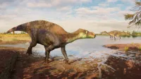 Una reconstrucción del dinosaurio iguanodonte Palodohadros Proudlovii caminando a través del barro a lo largo del borde de un cuerpo de agua poco profundo. A lo lejos, el gran dinosaurio