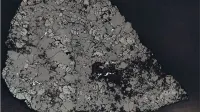 Una sección transversal microscópica de un espécimen de roca colocado sobre un fondo negro, que muestra una textura mineral muy fracturada, parecida a un mosaico en tonos de gris y negro