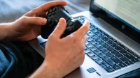 Person holds a gaming controller while interacting with a laptop, emphasizing modern gaming technology.