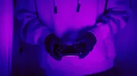 Una persona en una sudadera con capucha tiene un controlador de juego bajo una luz púrpura neón.