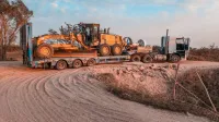 Un camión de carga transportando maquinaria pesada en un camino de tierra accidentado bajo un cielo despejado.