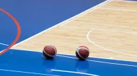 Two basketballs resting on a blue indoor basketball court with wooden flooring.