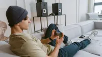 Joven casual jugando juegos móviles en el sofá con altavoces en el fondo.
