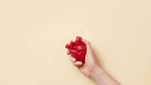 Un modelo de corazón anatómico rojo realista sostenido firmemente en una mano sobre un fondo beige.