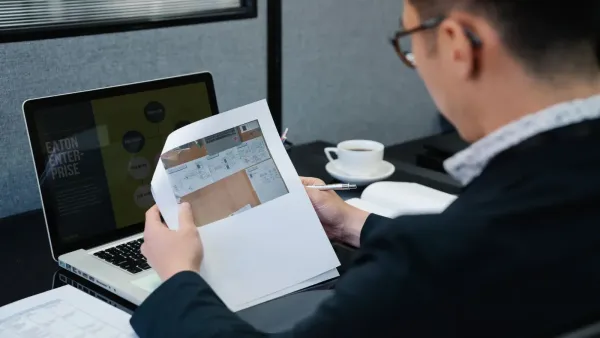 Un hombre en una oficina analizando documentos con una computadora portátil abierta cerca.
