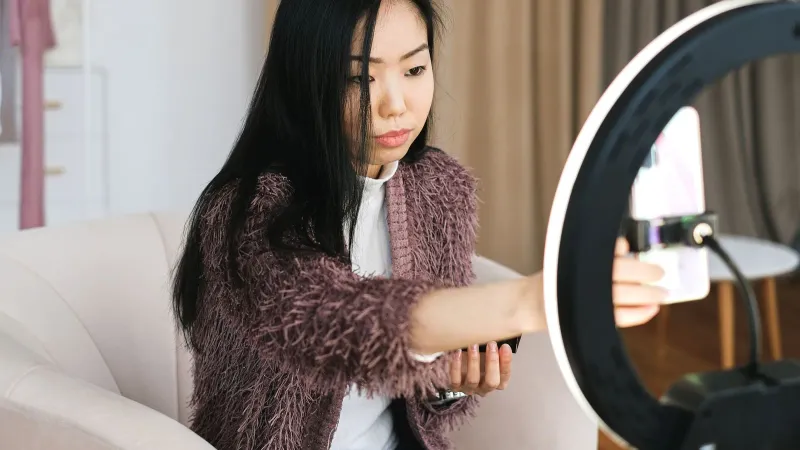 An Asian woman vlogs at home using a smartphone and ring light, perfect setup for influencers.