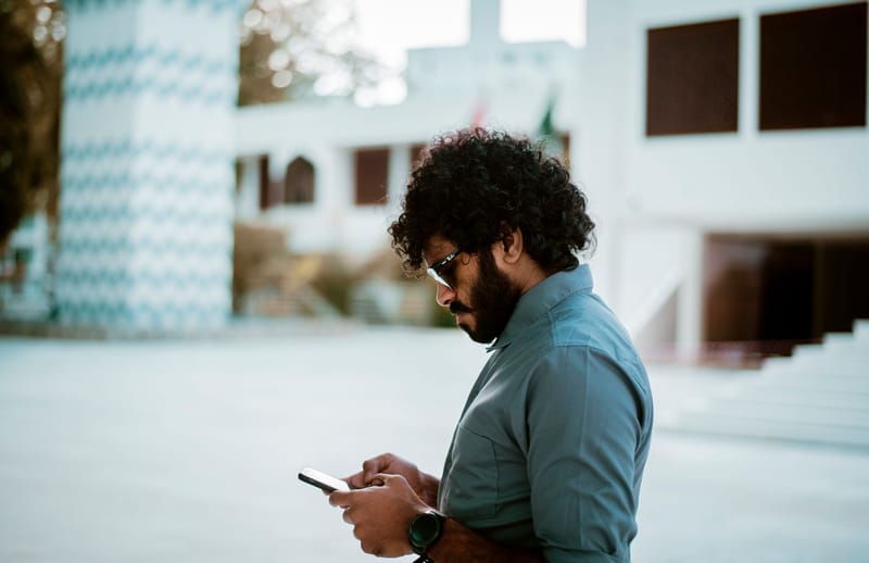 Side view of a curly-haired man with sunglasses using a smartphone in a sunny outdoor setting.