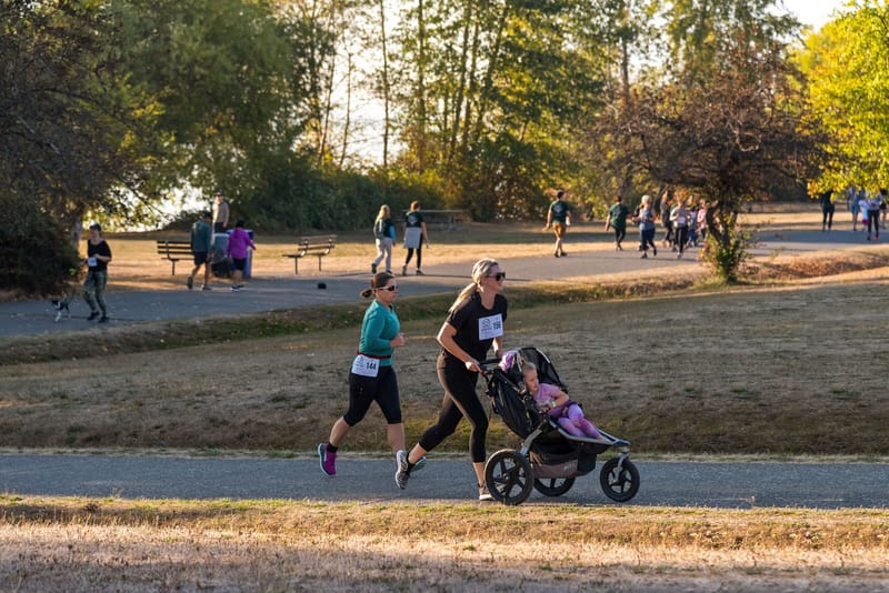 A woman pushing a stroller during a marathon in a sunny park setting, surrounded by fellow runners.