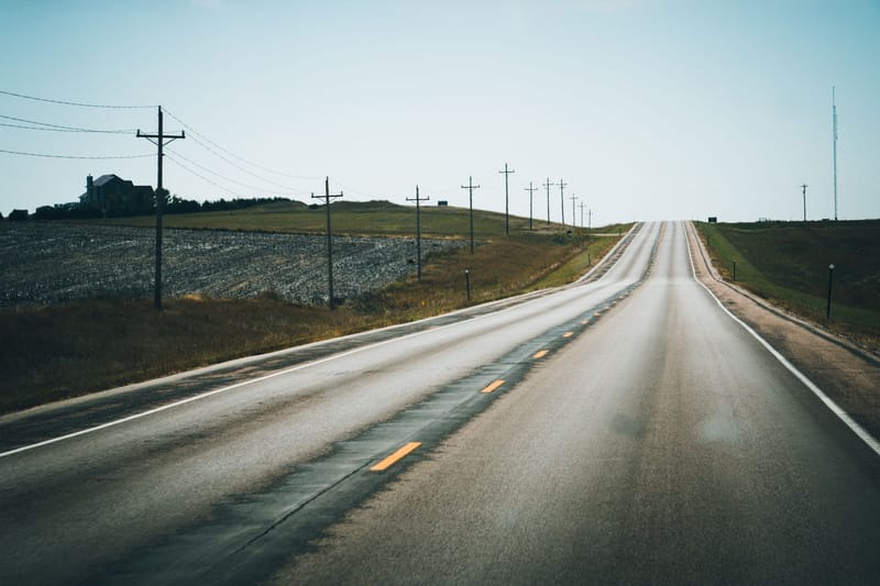 A peaceful rural highway stretching into the distance, flanked by telephone poles and open fields under a clear summer sky.