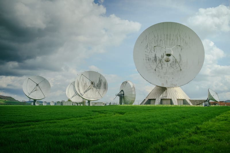 Array of large satellite dishes in a lush green field under a cloudy sky.
