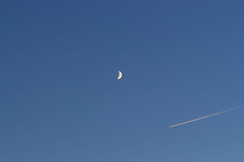 A crescent moon and airplane contrail against a clear blue sky, creating a serene contrast.