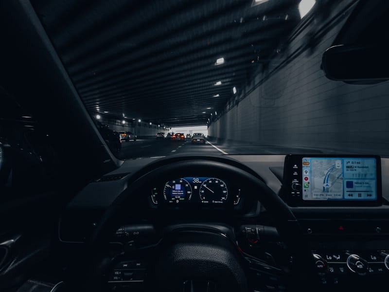 View from car dashboard driving through an illuminated city tunnel at night.