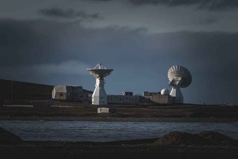 A remote satellite station with large dishes stands under a moody dark sky by the water.