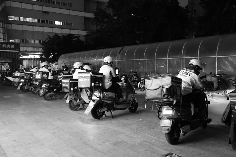 Food delivery drivers on scooters gather at night for dispatch in an urban setting.