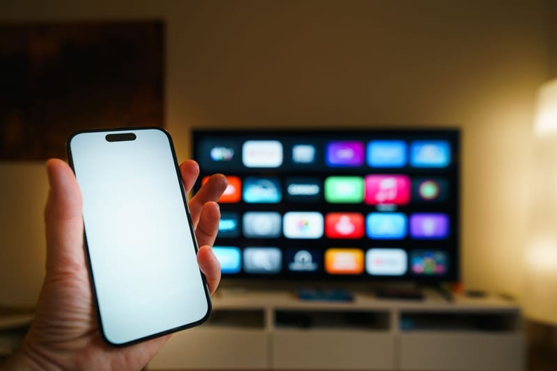 A smartphone in focus with a smart TV displaying apps in a cozy living room.