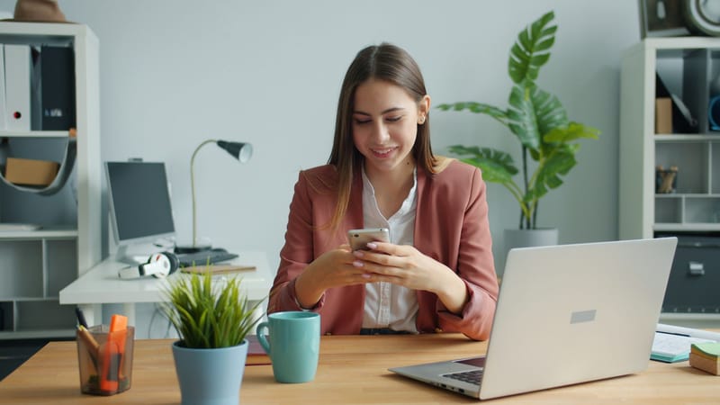 Smiling businesswoman checking phone, seated at desk with laptop and plants.