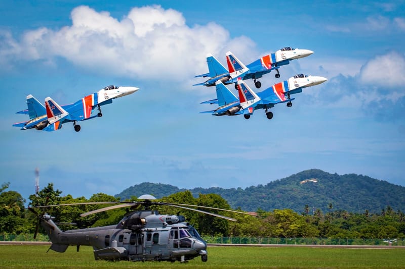 Three aerobatic jets performing a formation flight above a helicopter on a clear sunny day.