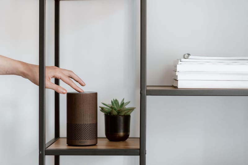 A hand interacts with a smart speaker on a minimalist shelf, featuring a plant and stacked books.