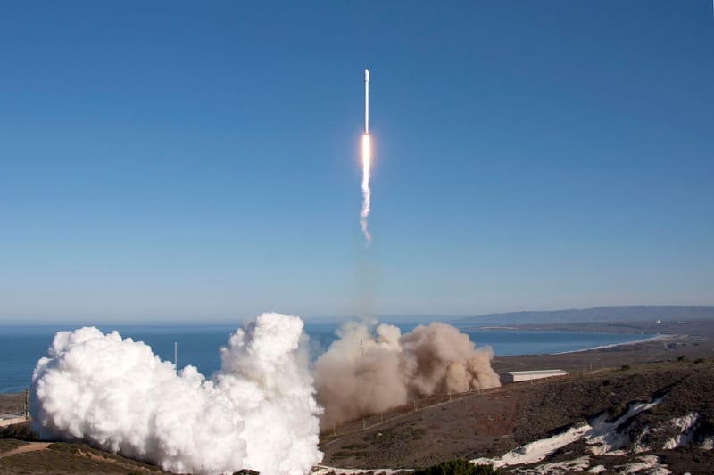 A rocket launches over the ocean, leaving behind smoke and fire trails. Scenic backdrop with clear skies.