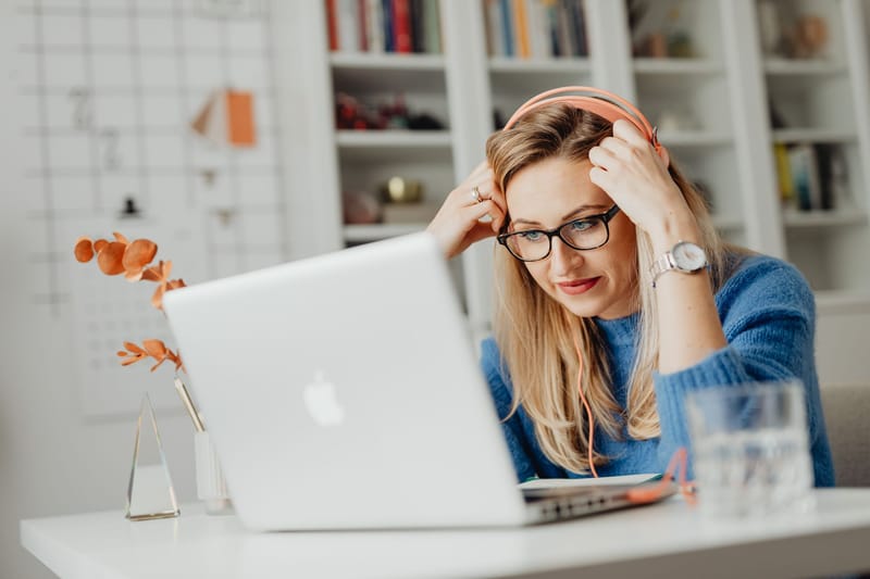 Blond woman wearing eyeglasses and headphones, working on a laptop indoors at a desk.