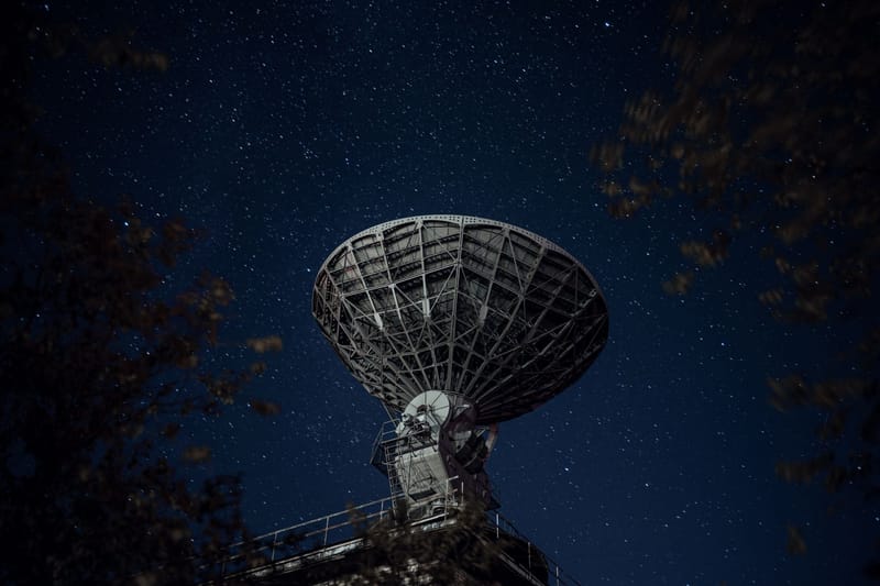 From below of blue starry sky over radio telescope and trees with leaves