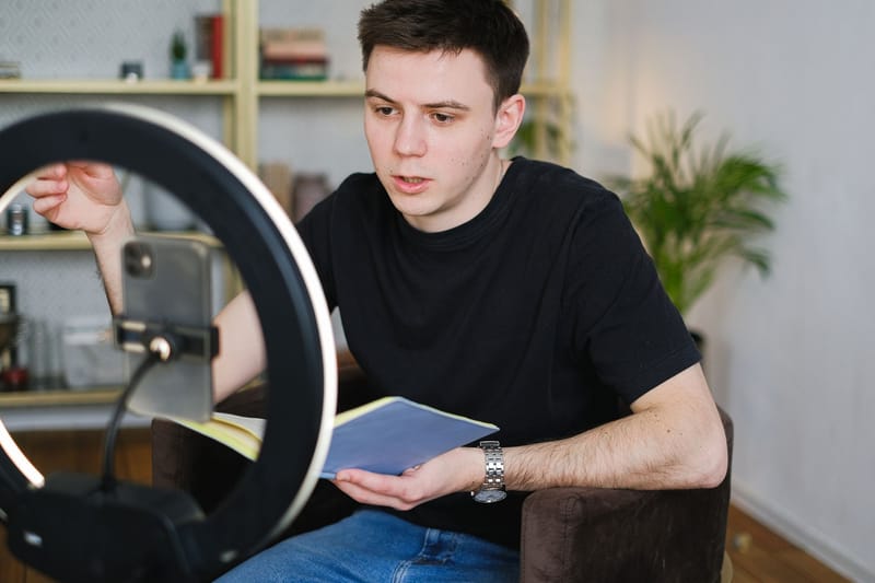 Adult male recording video content indoors, using a smartphone and ring light, holding a notebook.
