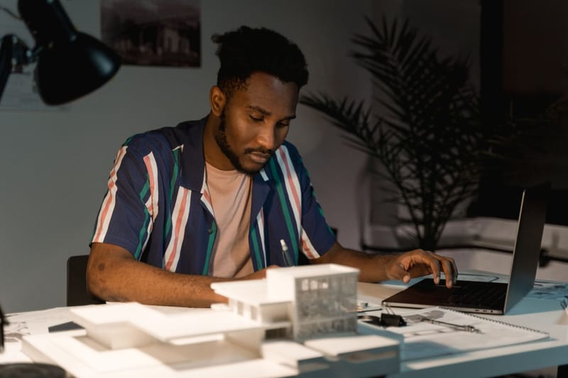 An architect working late at night in an office, using a laptop and surrounded by building models.