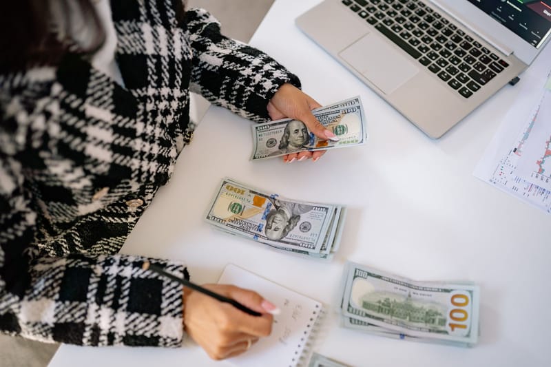 A woman in a plaid coat counts American dollars at a desk with a notebook and laptop.