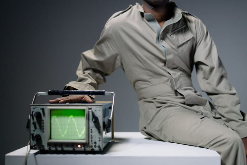 A person in coveralls sits beside an oscilloscope displaying a waveform in a studio setting.