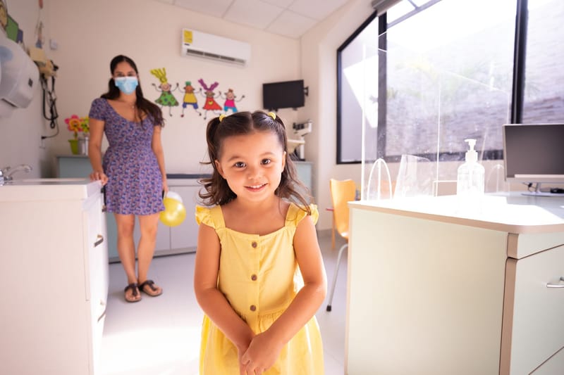 A cheerful child in a yellow dress at a pediatric clinic with doctor in background.