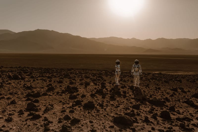Astronauts in space suits explore a rocky, desert-like terrain resembling Mars at sunset.