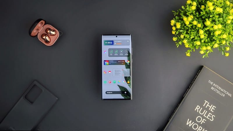 Top-down view of a smartphone, earbuds, book, and plant on a dark surface.