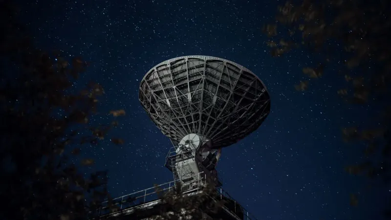 From below of blue starry sky over radio telescope and trees with leaves