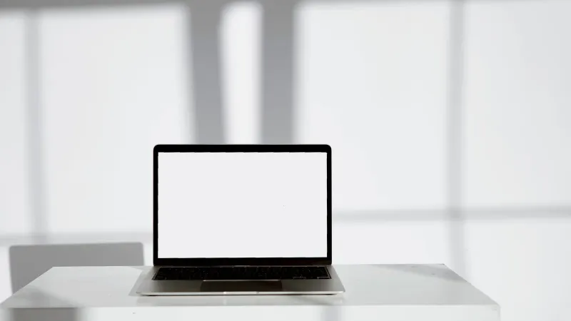 Blank laptop screen on a modern white desk in a bright, sunlit room.