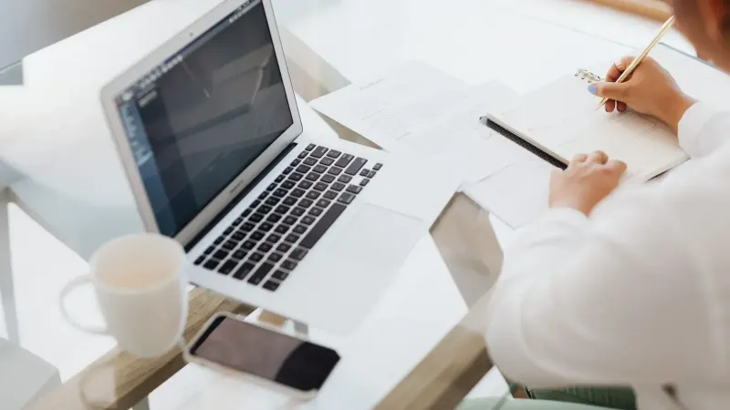 Persona que trabaja en una mesa de vidrio con una computadora portátil, cuaderno y taza de café.