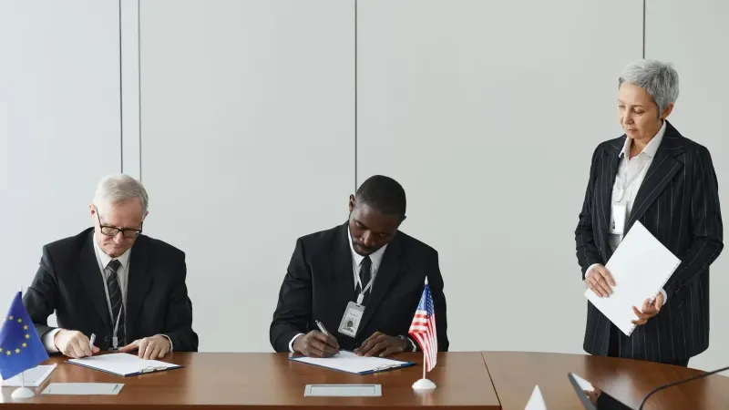 Executives signing international agreement with EU and US flags displayed on a wooden table.