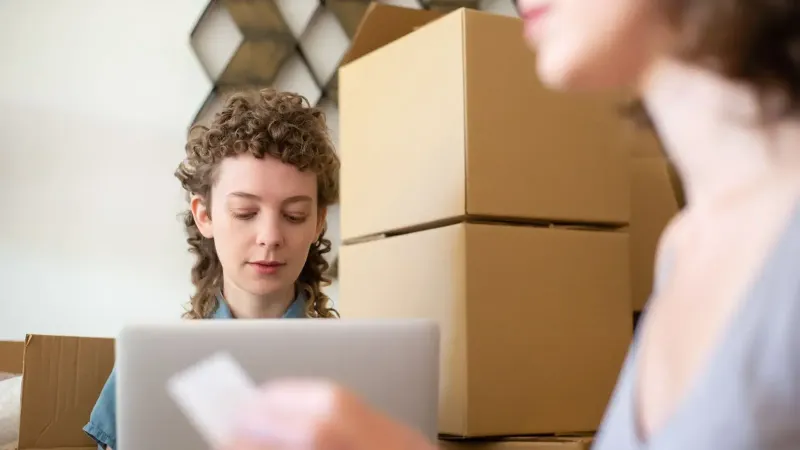 Women managing a small business surrounded by cardboard boxes and using a laptop for online orders.