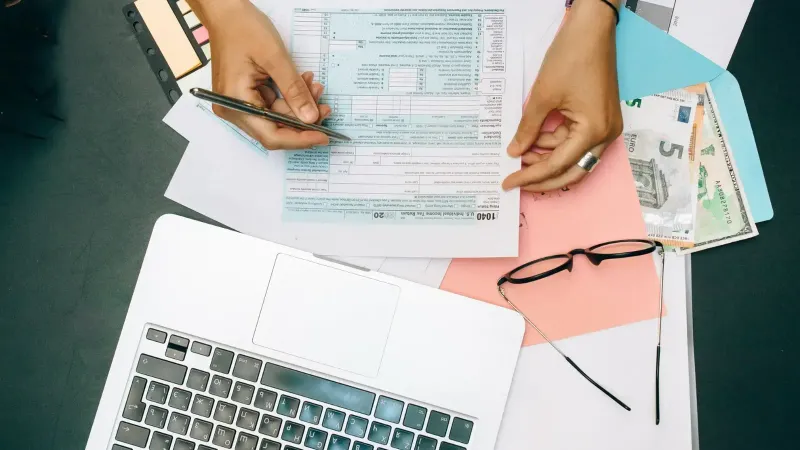 Manos escribiendo documentos fiscales con computadora portátil, anteojos y moneda en el escritorio.