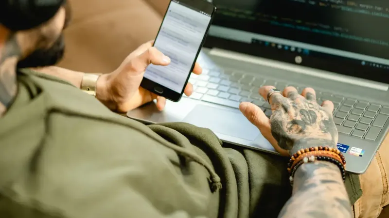Una persona tatuada se dedica a la codificación en una computadora portátil mientras revisa un teléfono inteligente.