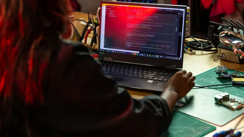 A software developer writing code on a laptop at a cluttered workbench with electronic tools.
